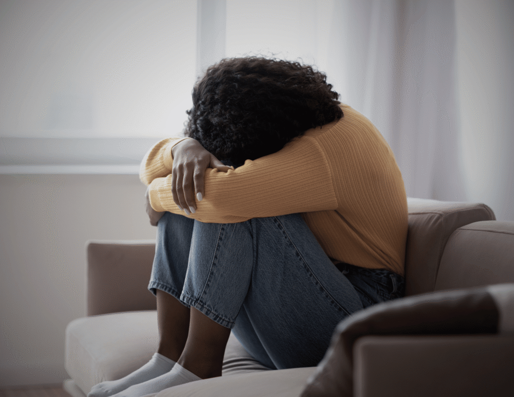 Unhealthy ways people deal with depression — a young Black woman sitting alone on a couch with her head buried in her knees