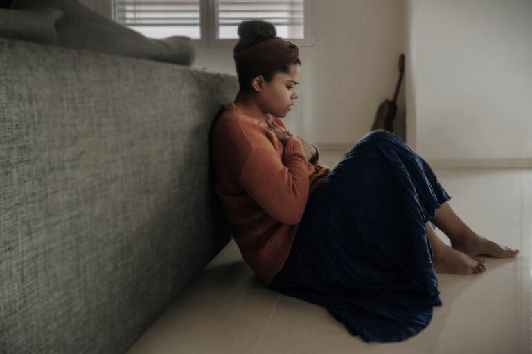 Woman sitting on the floor with her hand on her chest, leaning against a bed, symbolizing anxiety or emotional distress.