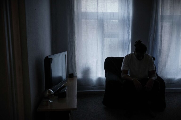 A man sits alone in a dimly lit living room, his face hidden in the shadows. A faint glow from the television is the only light in the room, casting a somber and isolated mood.