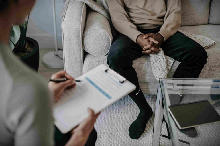 A man sits on a couch in a therapy room, talking to a female therapist who is taking notes. Their faces are not visible, creating an anonymous and relatable scene that focuses on the therapy process.