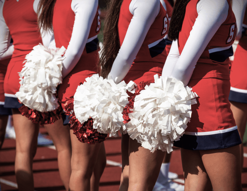 A row of cheerleaders in red and white uniforms holding white and red pom-poms, photographed from the waist down on a sports field