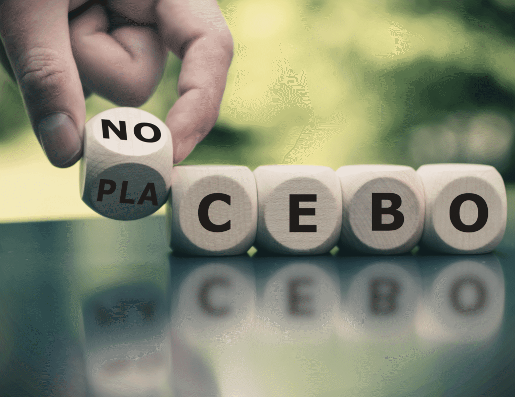 A hand switching a wooden dice from reading "PLA" to "NO" in front of three other dice spelling "CEBO," together forming the word "Nocebo" instead of "Placebo," reflected on a glass surface with a green blurred background