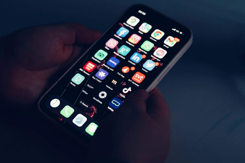 A hand holding a smartphone in a dark room, its screen brightly lit with a full grid of app icons including Facebook, Instagram, TikTok, LinkedIn, and Gmail