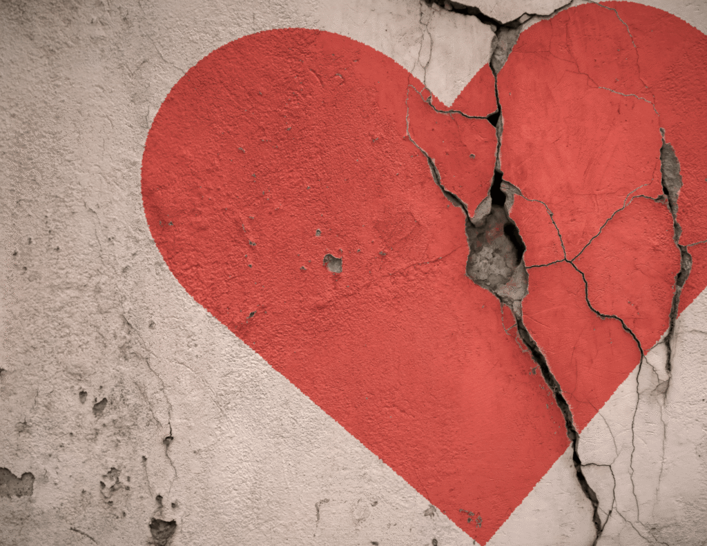 A red heart painted on a cracked and crumbling concrete wall, with deep fractures splitting through the right side of the heart, symbolizing heartbreak and emotional pain.