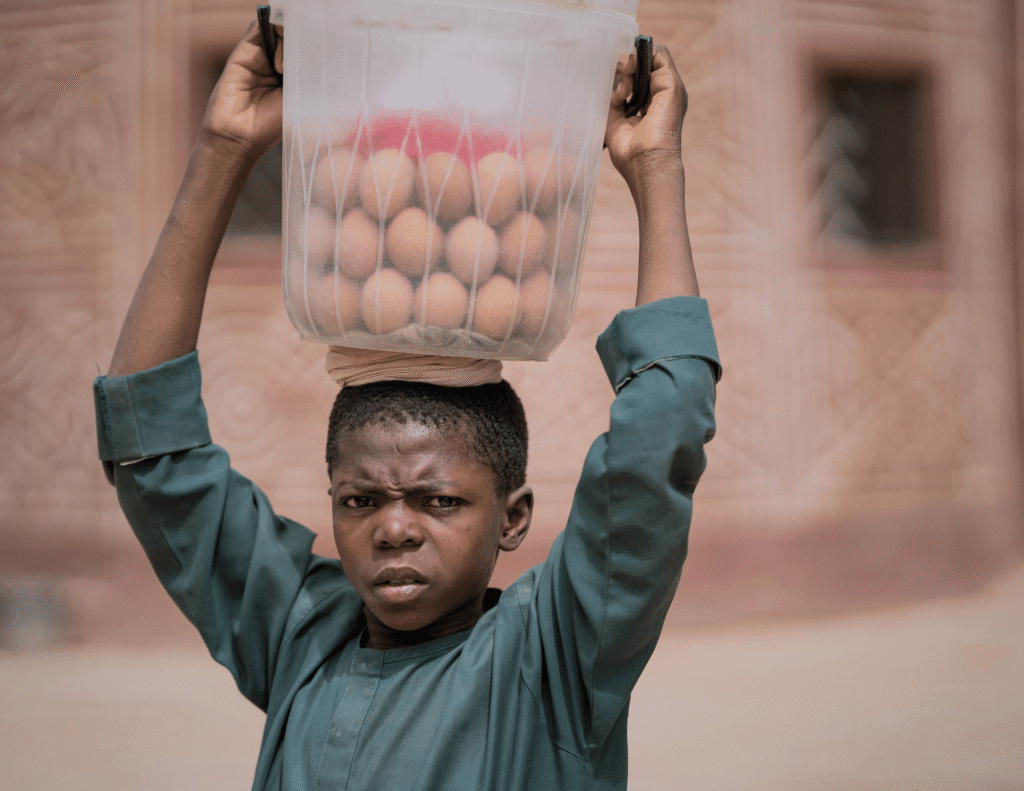 A young African boy carrying a large container of eggs on his head, visibly strained, representing child labour and firstborn responsibilities in African homes