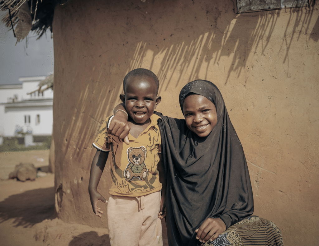 A smiling older African girl in a hijab posing with her younger brother outside a mud house, representing the sibling caretaker role of firstborns in African families