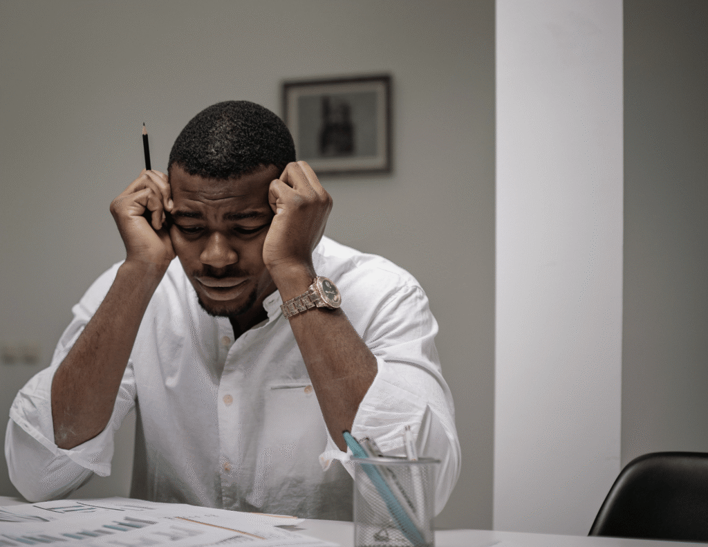 Young Nigerian man sitting at a desk with his head in his hands, overwhelmed by academic and work pressure