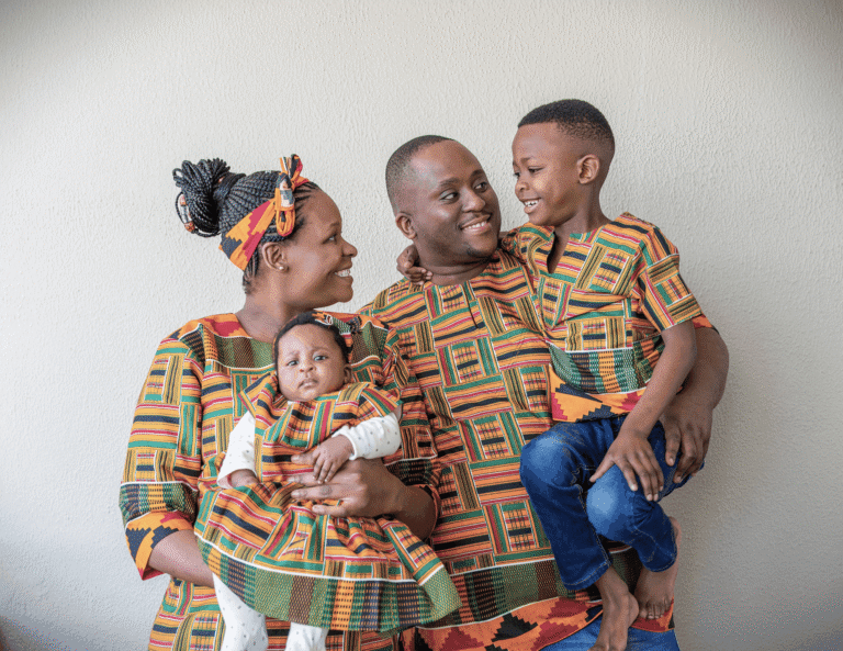 A happy African family in matching kente outfits — mother holding a newborn, father carrying the firstborn son — symbolising the cultural weight and expectations placed on firstborn children in African homes