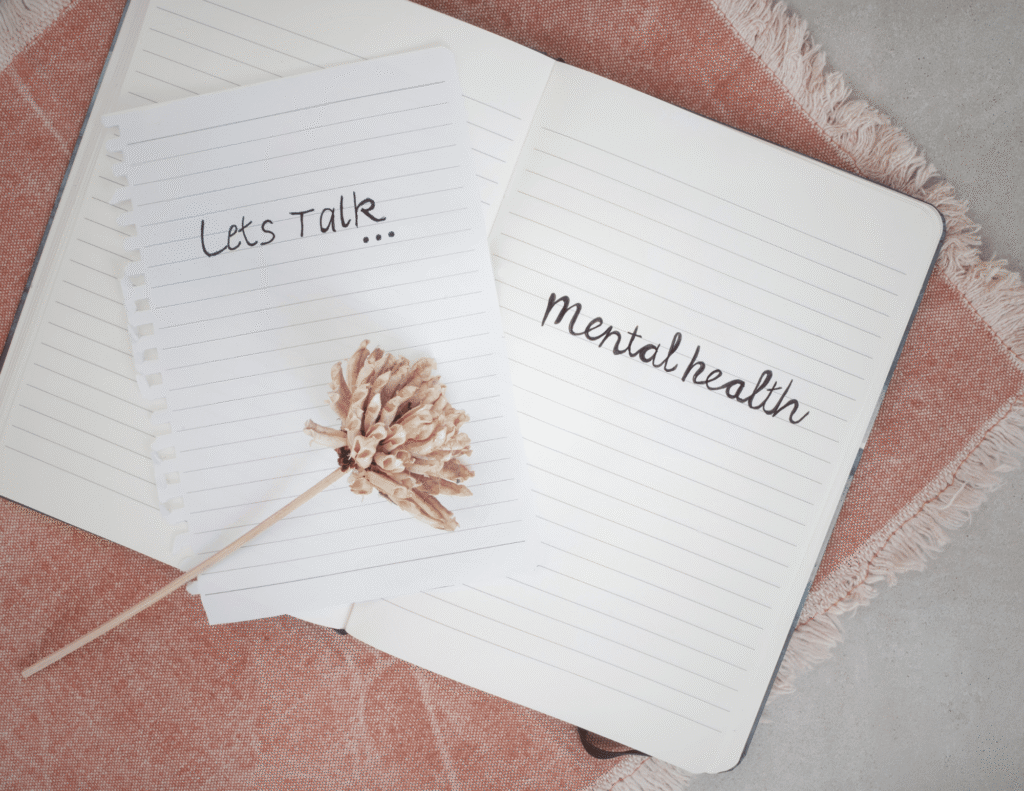 Mental Health Awareness Month Two open notebooks with the words "Let's Talk" and "mental health" written by hand, placed on a pink textured cloth with a dried flower between them